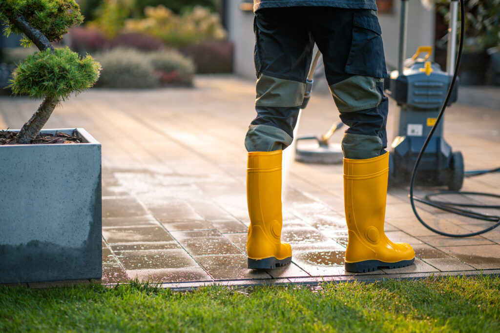 A person stands on a patio, wearing yellow waterproof boots, operating a pressure washer while water sprays onto the tiles, enhancing the garden's appearance.
